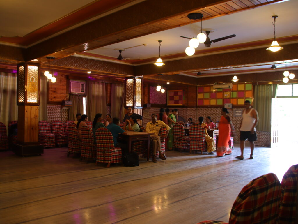 Dining hall with wooden ceiling, decorative lighting, and marble flooring. People are seated around tables covered with checkered cloth chairs, while others stand and walk around. The room has air conditioning units, patterned walls, and large windows with curtains.